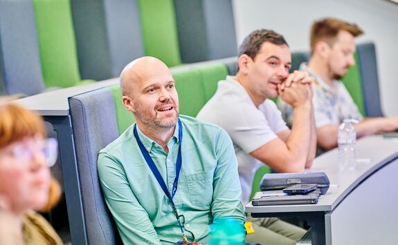 Group of people seated in a lecture hall with tiered green and grey cushioned seating. One person in the foreground wears a light green shirt and a lanyard.